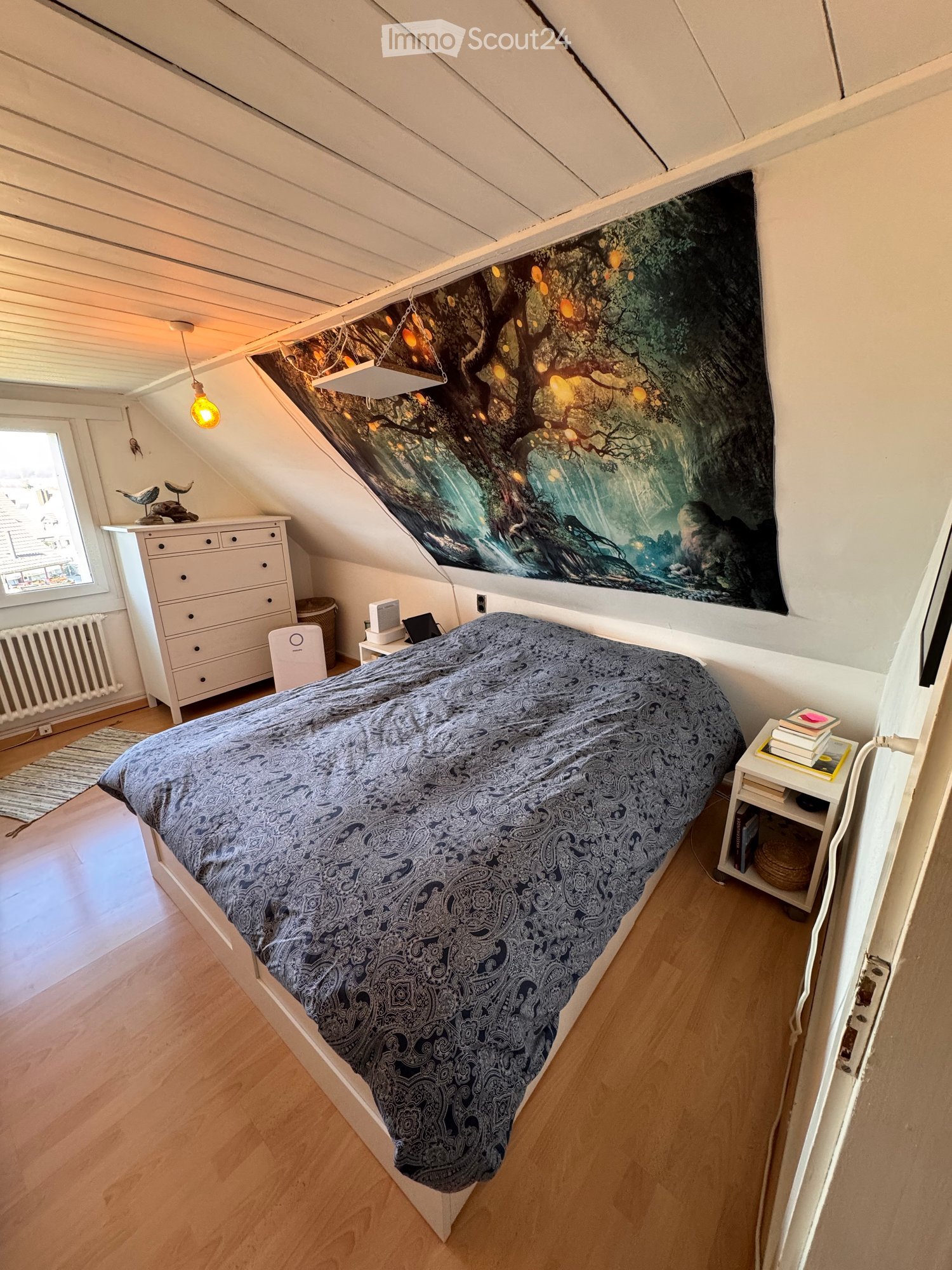 Cozy attic bedroom with wooden floors, slanted ceilings, a large bed with patterned bedding, a nightstand with books, a drawer unit, and a large wall mural.