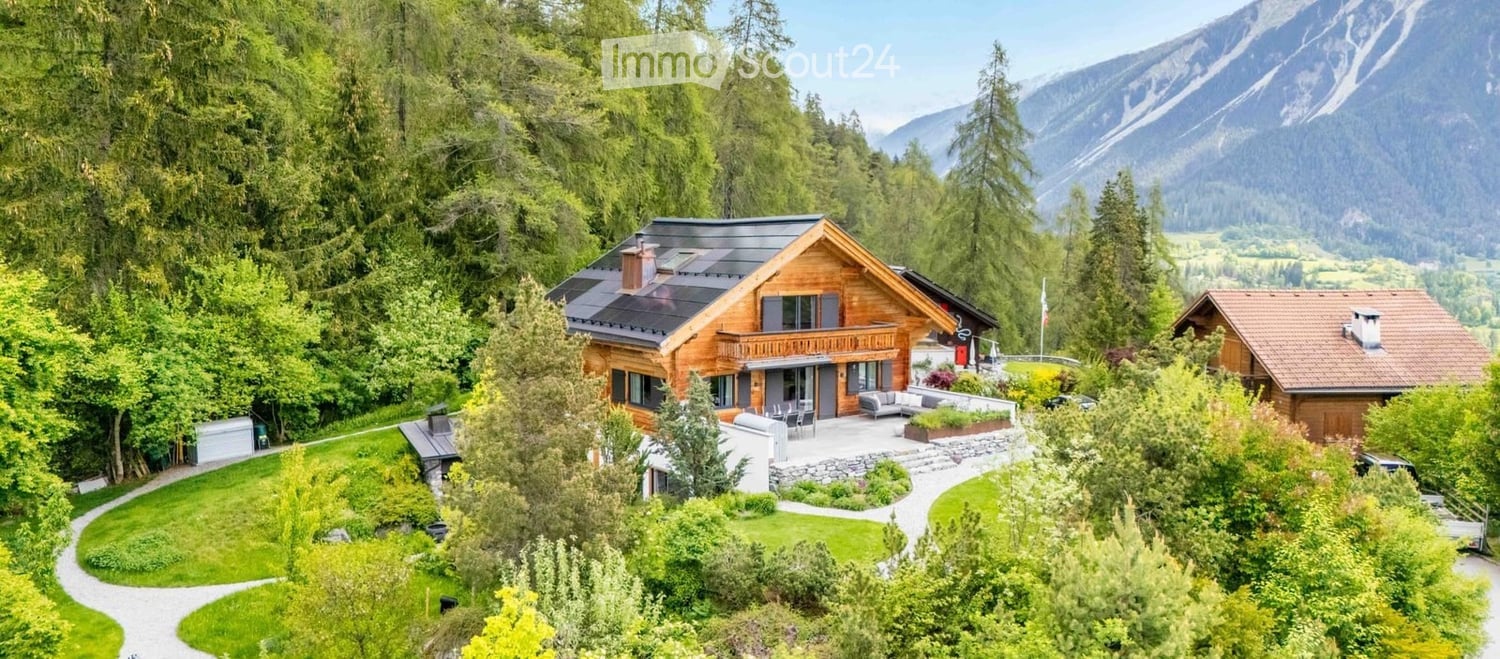 A large, modern wooden house with a balcony, surrounded by lush greenery and mountains in the background.