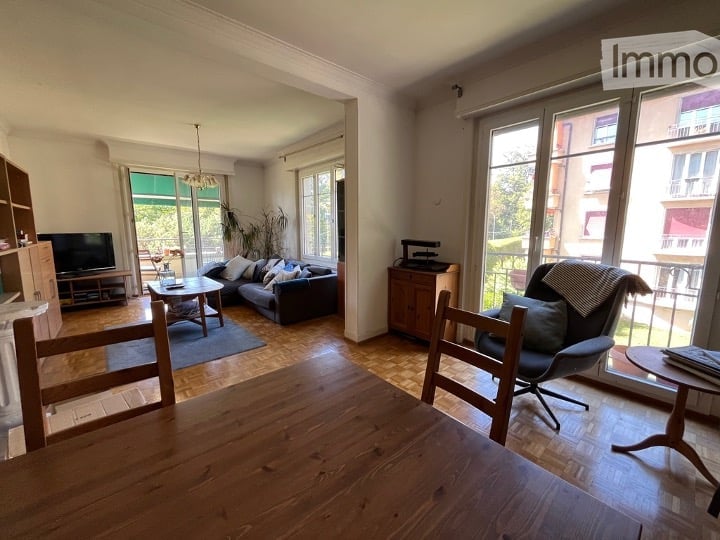 Bright living room with large windows and a view, featuring a sectional sofa, coffee table, dining table, chairs, TV, and wooden flooring.