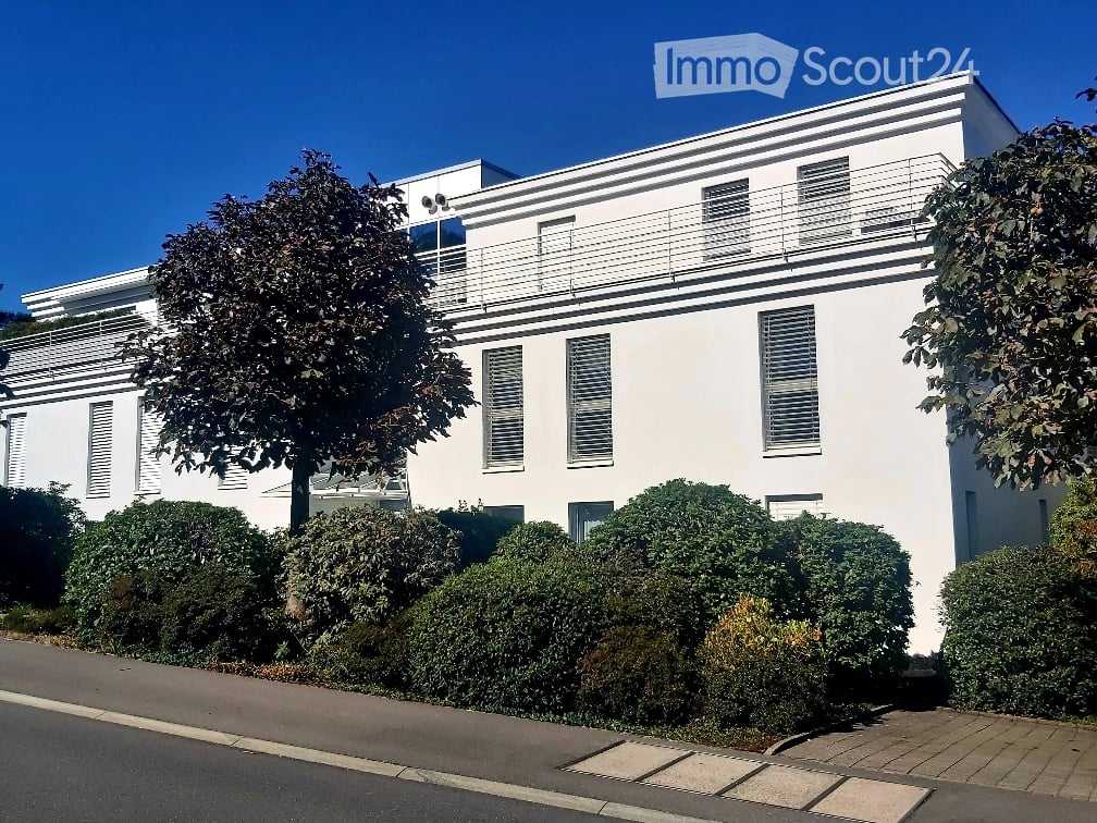 Modern two-story white building with large windows, balconies, and trimmed hedges in front.