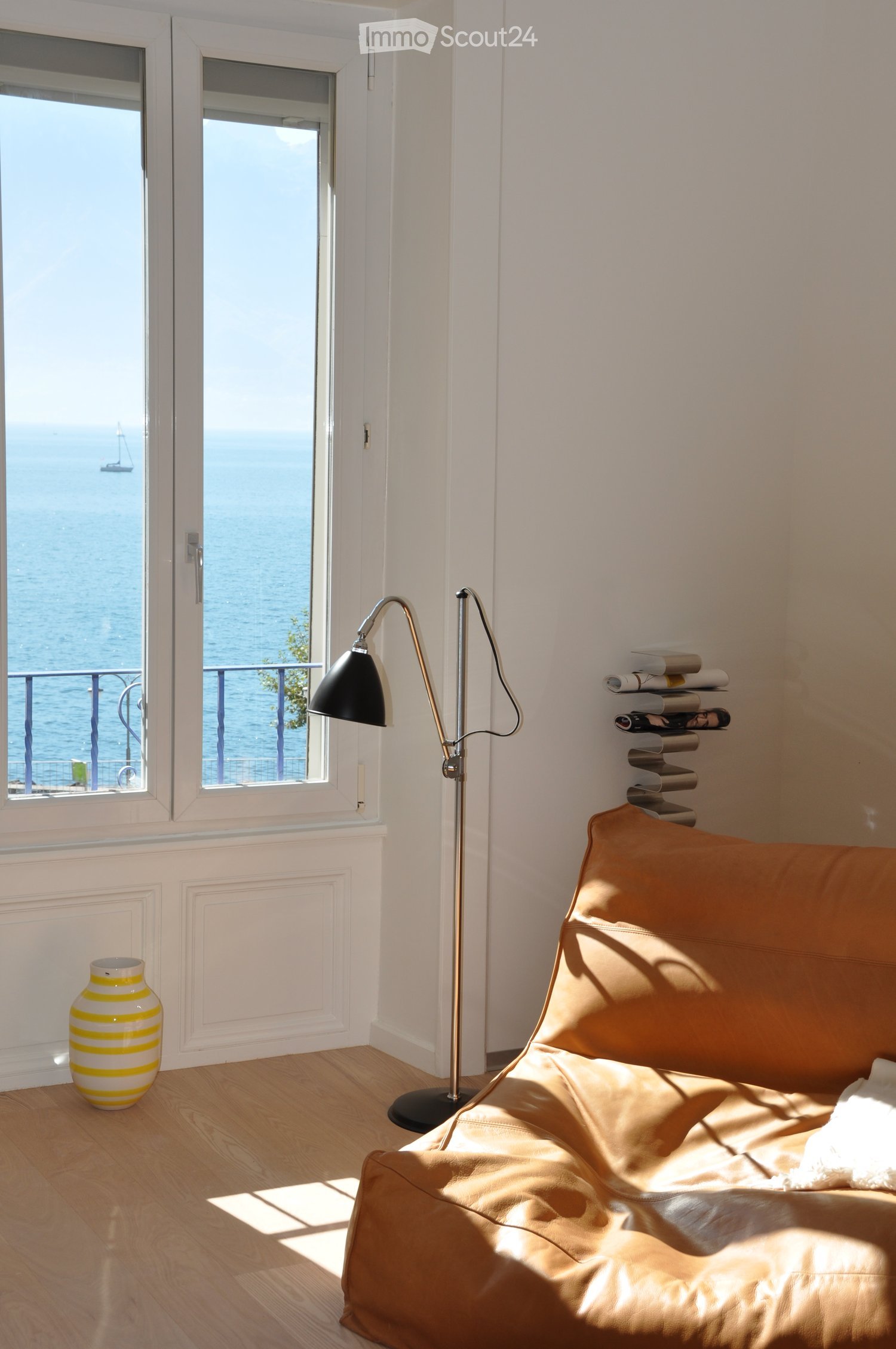 Bright living room with a tan leather sofa, floor lamp, decorative vase, and magazine rack, overlooking the sea through large windows.