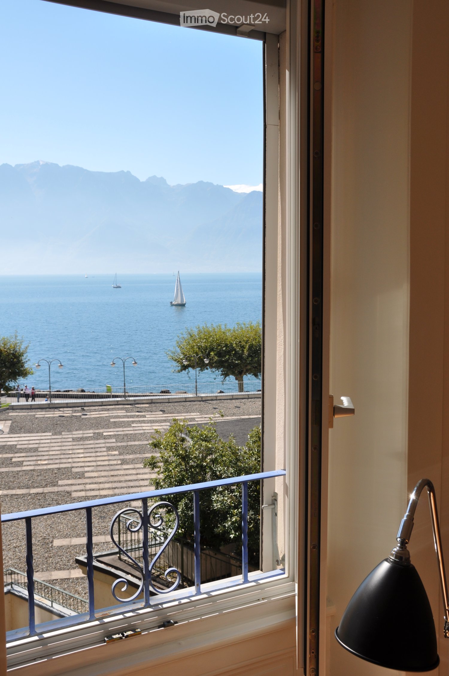 Balcony with a view of a lake, sailboats, and mountains, featuring a decorative metal railing and a modern lamp inside the room.