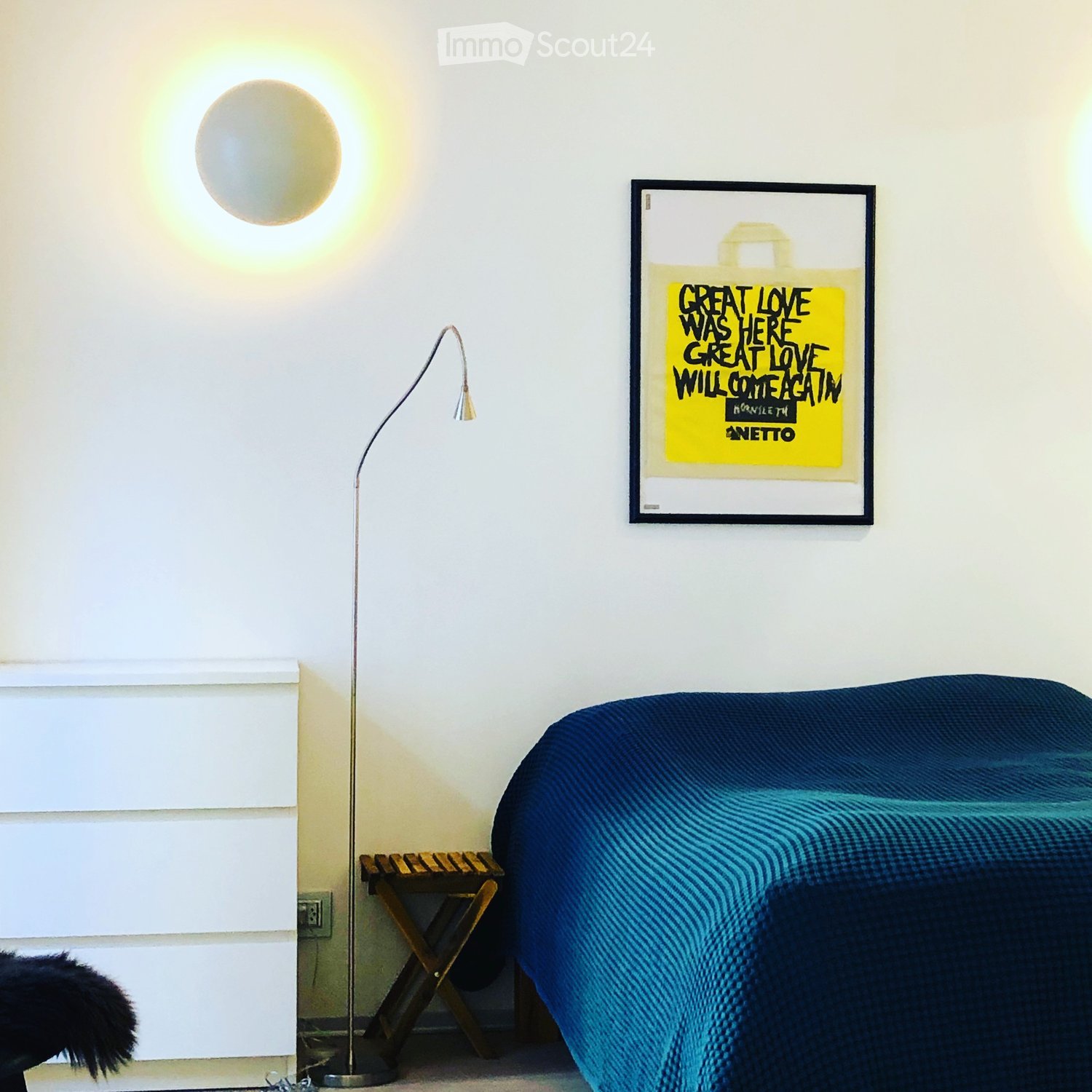 Bedroom with a blue bedspread, a wooden stool, a floor lamp, and a framed picture on the wall.