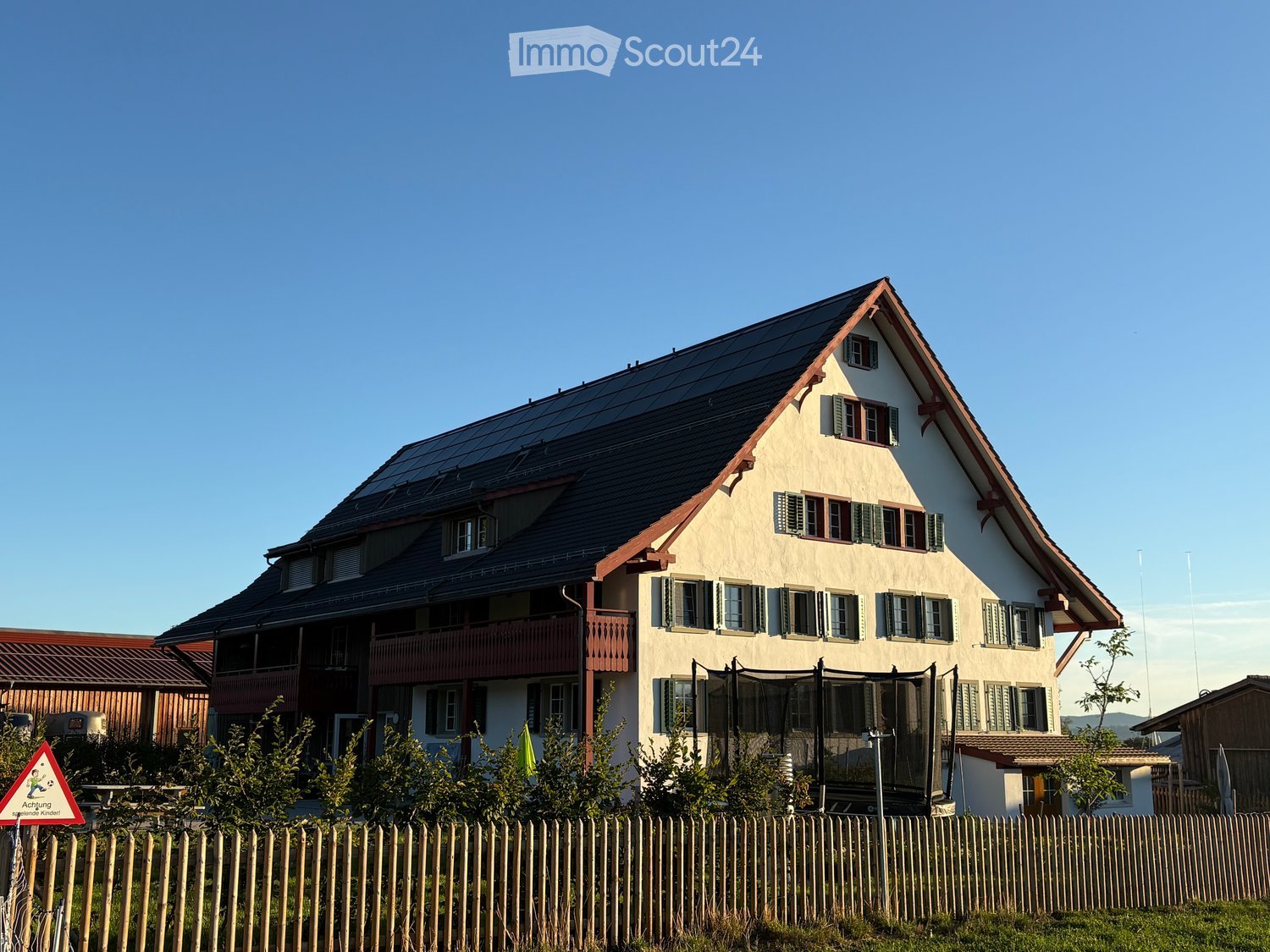 Two-story house with a pitched roof, multiple windows, and a balcony on the upper floor. The house has solar panels on the roof and is surrounded by a wooden fence and greenery.
