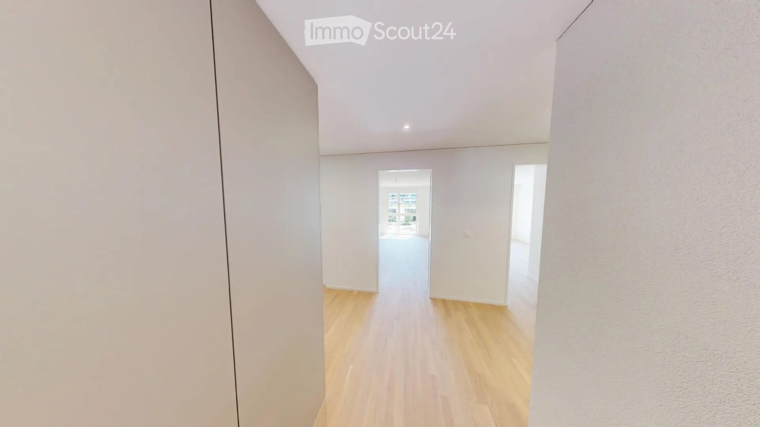 Empty hallway with light wooden flooring, white walls, and recessed lighting. Two open doorways lead to other rooms.