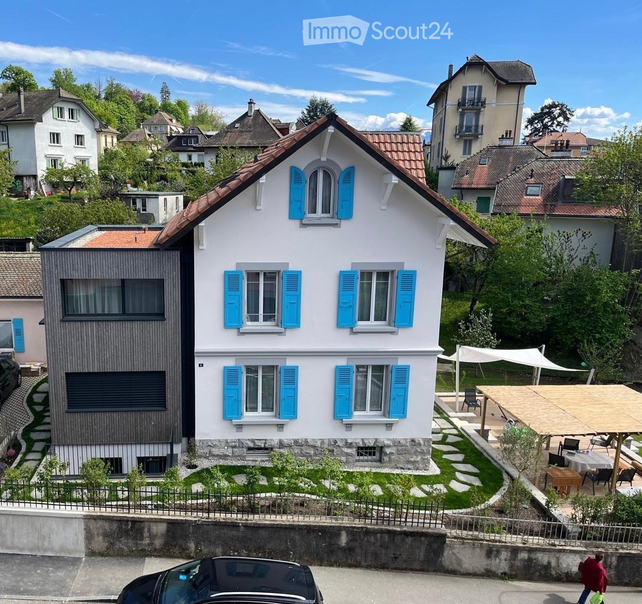 Two-story house with white walls, blue shutters, and a red-tiled roof. The house features a modern extension with dark wooden panels and large windows. The front yard includes a small garden with stepping stones and a covered outdoor seating area with tab