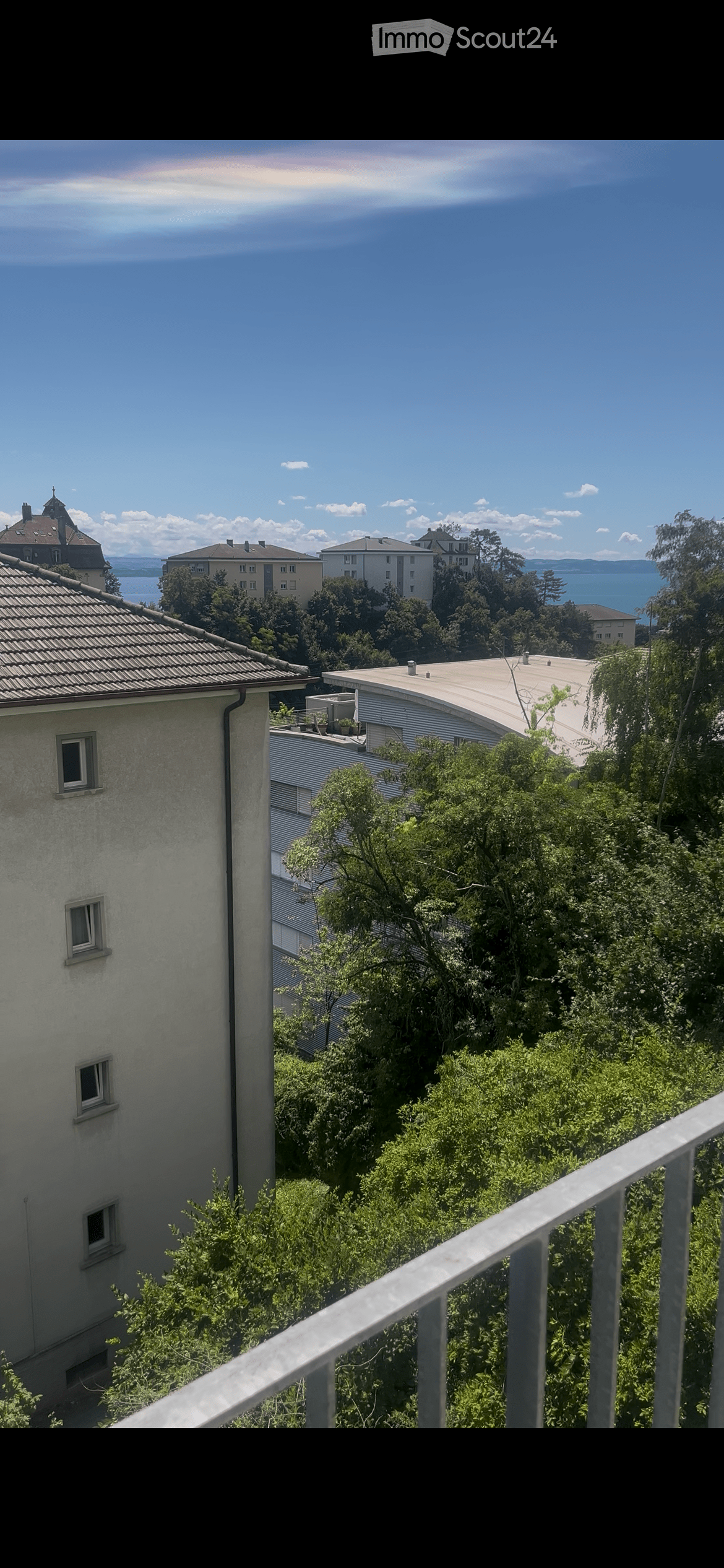 Balcony overlooking a lush green area with trees, featuring a metal railing and a view of neighboring buildings and a body of water in the distance.