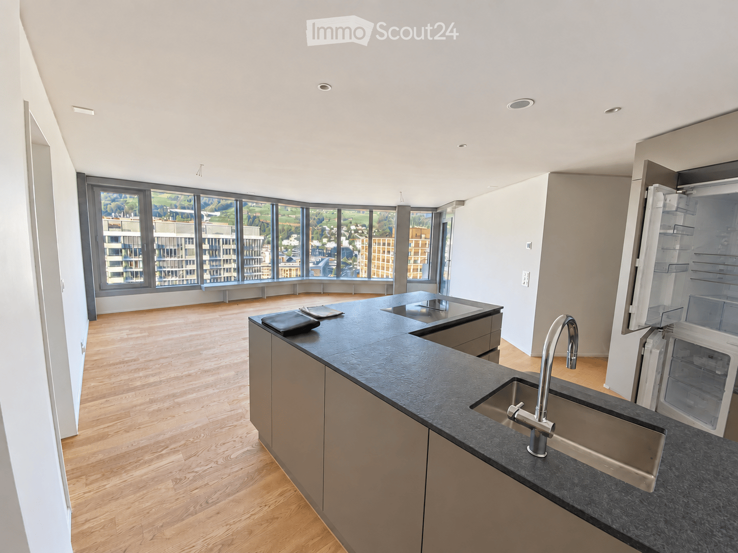 Modern kitchen with wooden flooring, large windows, stainless steel sink, faucet, and refrigerator.