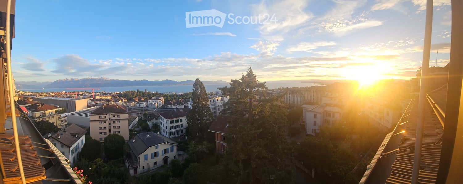Balcony with a panoramic view of a cityscape at sunset, featuring buildings, trees, and distant mountains and water.