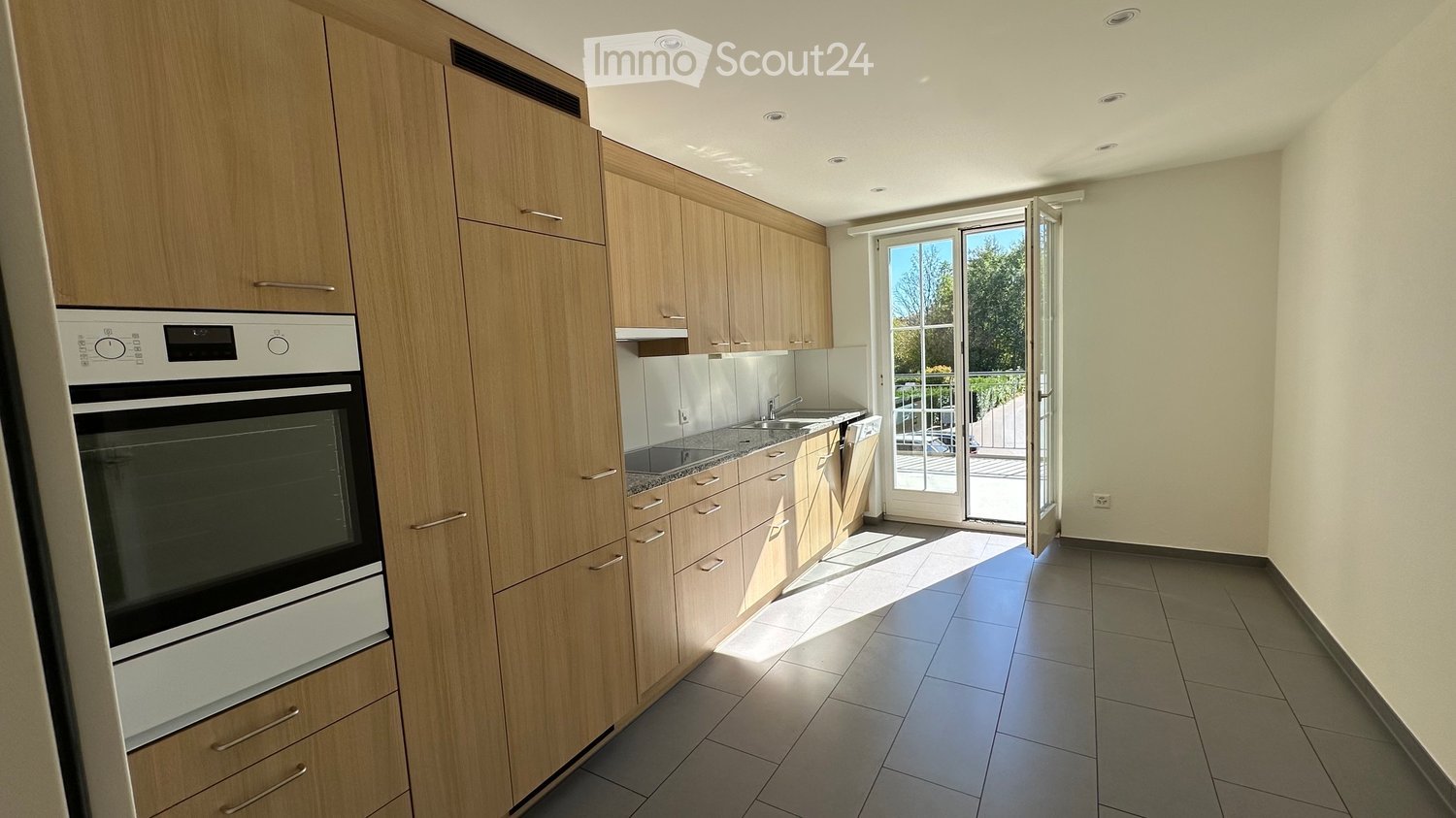 Modern kitchen with light wooden cabinets, stainless steel oven, sink, and faucet. Large sliding glass door leads to a balcony or outdoor area.