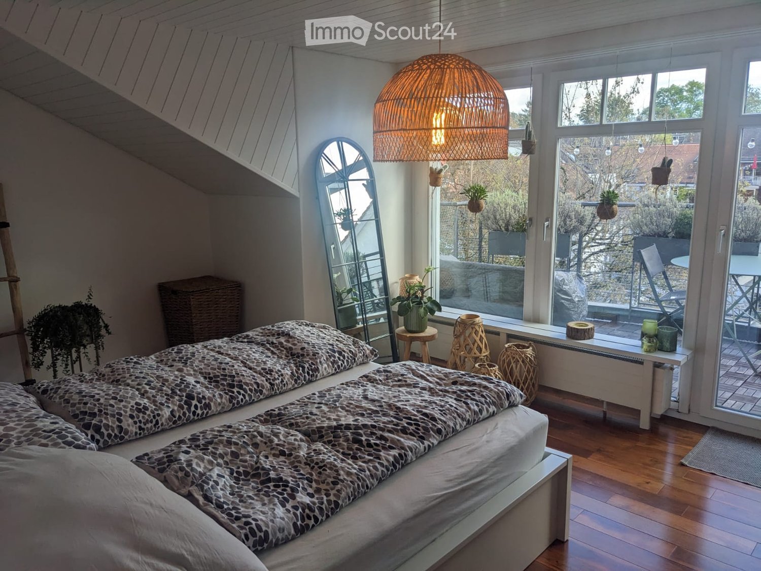 Cozy bedroom with a large bed, patterned bedding, wooden floor, large windows with a view, and decorative items including a wicker lamp, mirror, and potted plants.