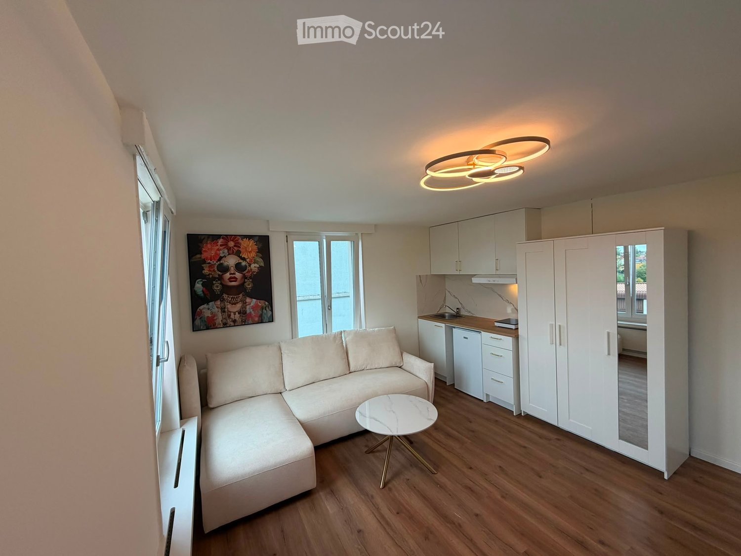 Modern, open-plan living and kitchen area with a light-colored sofa, coffee table, kitchen cabinets, sink, fridge, and a large painting on the wall.