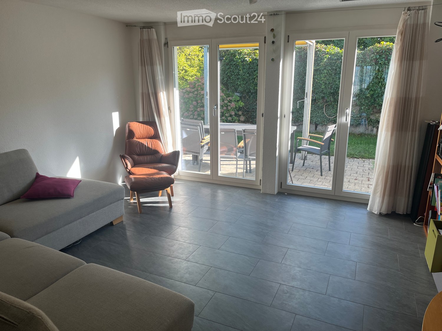 Bright living room with large sliding glass doors leading to a patio, featuring a grey sofa, brown leather chair with ottoman, and outdoor furniture visible through the doors.