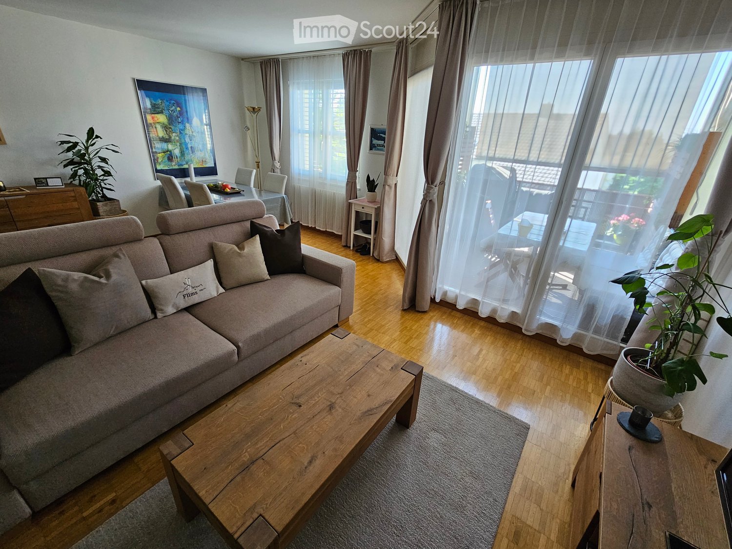 Modern living room with a grey sofa, wooden coffee table, side table, dining area with chairs, and large windows with sheer curtains.
