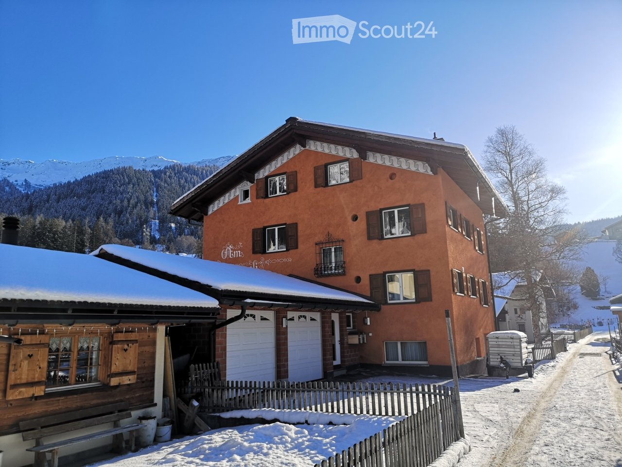 A two-story house with a red-orange facade, multiple windows, and a snow-covered roof, situated in a snowy mountain landscape.