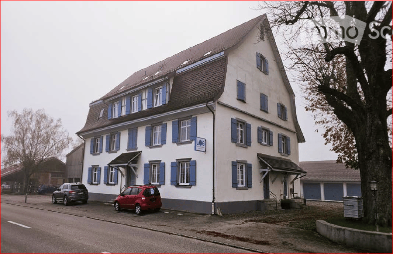 Two-story building with white walls, blue shutters, and a brown roof. Two cars are parked in front of the building.