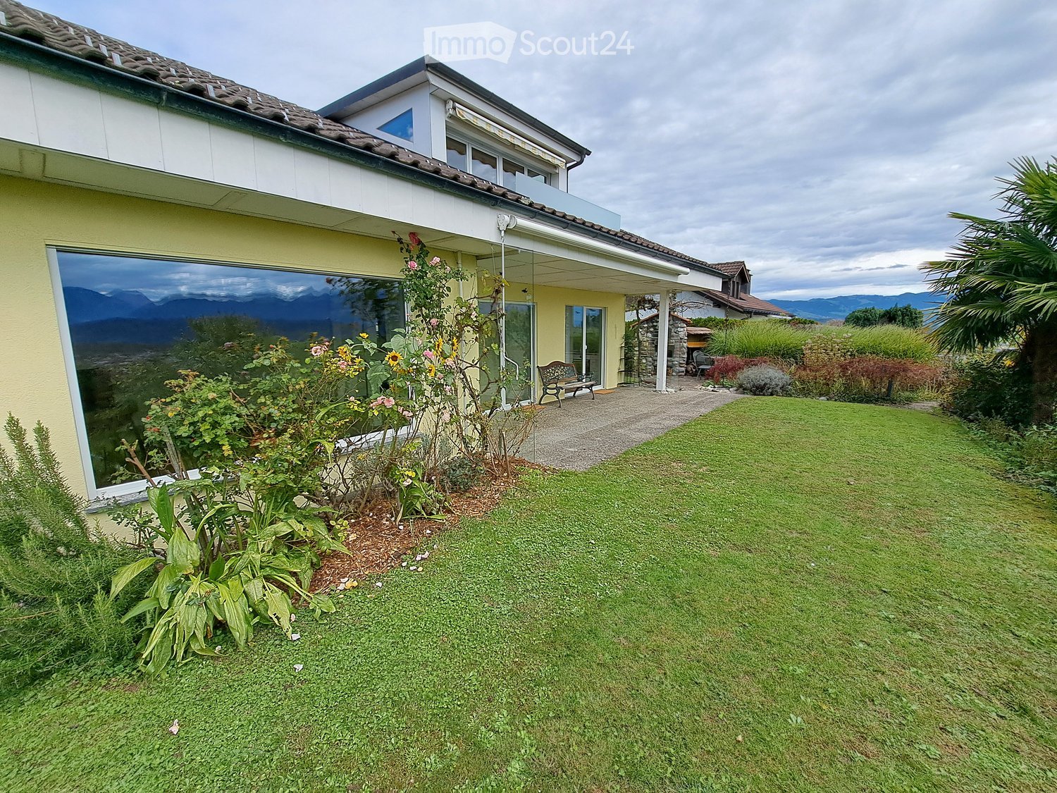 Well-maintained garden with a lawn, flower beds, and a bench near the house entrance. The house has large windows and a tiled roof, with a scenic mountain view in the background.