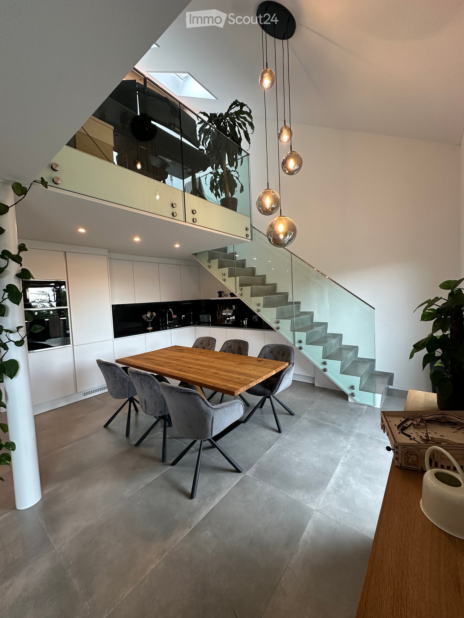 Modern open-plan dining and kitchen area with a wooden dining table, grey chairs, white cabinets, and a staircase leading to an upper level.