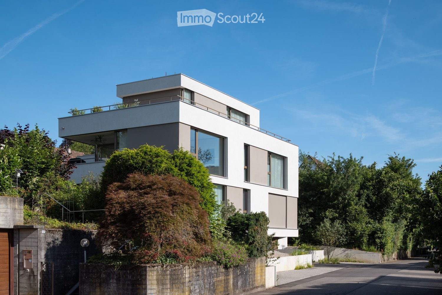 Modern two-story house with white exterior, large windows, and a raised ground floor, surrounded by greenery and trees.