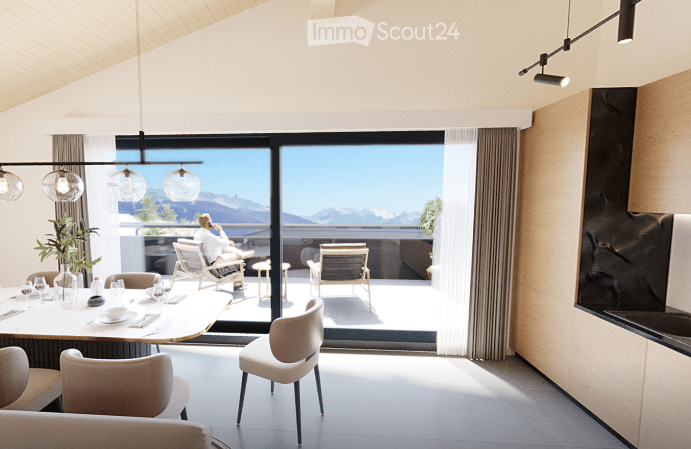 Modern dining area with a round table, chairs, and a view of mountains through large windows. There is a sofa and a plant in the foreground, and a person is sitting on a chair outside on the balcony.