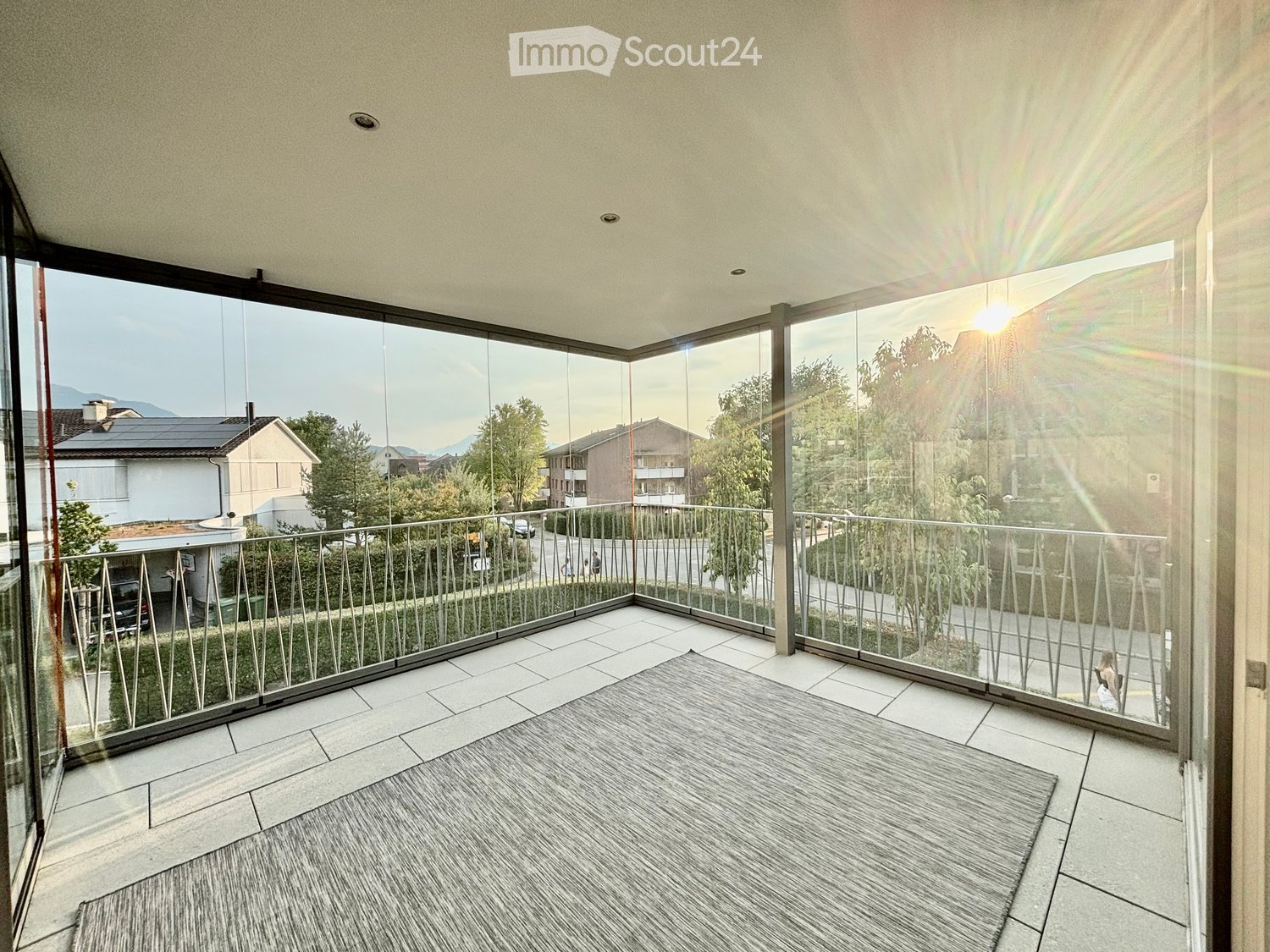 Modern balcony with glass railings, tiled floor, and a view of the surrounding neighborhood and mountains.