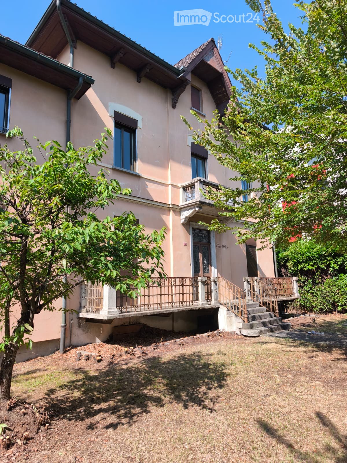 Two-story house with beige walls, multiple windows, and a small balcony with metal railing. The house has a staircase leading up to the entrance and is surrounded by trees and grass.
