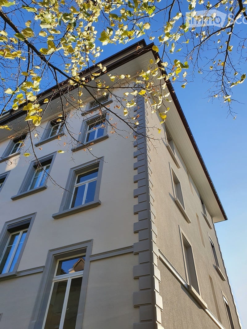 Four-story residential building with light beige facade, multiple windows, and decorative vertical siding. Overhanging roof and tree branches visible in foreground.