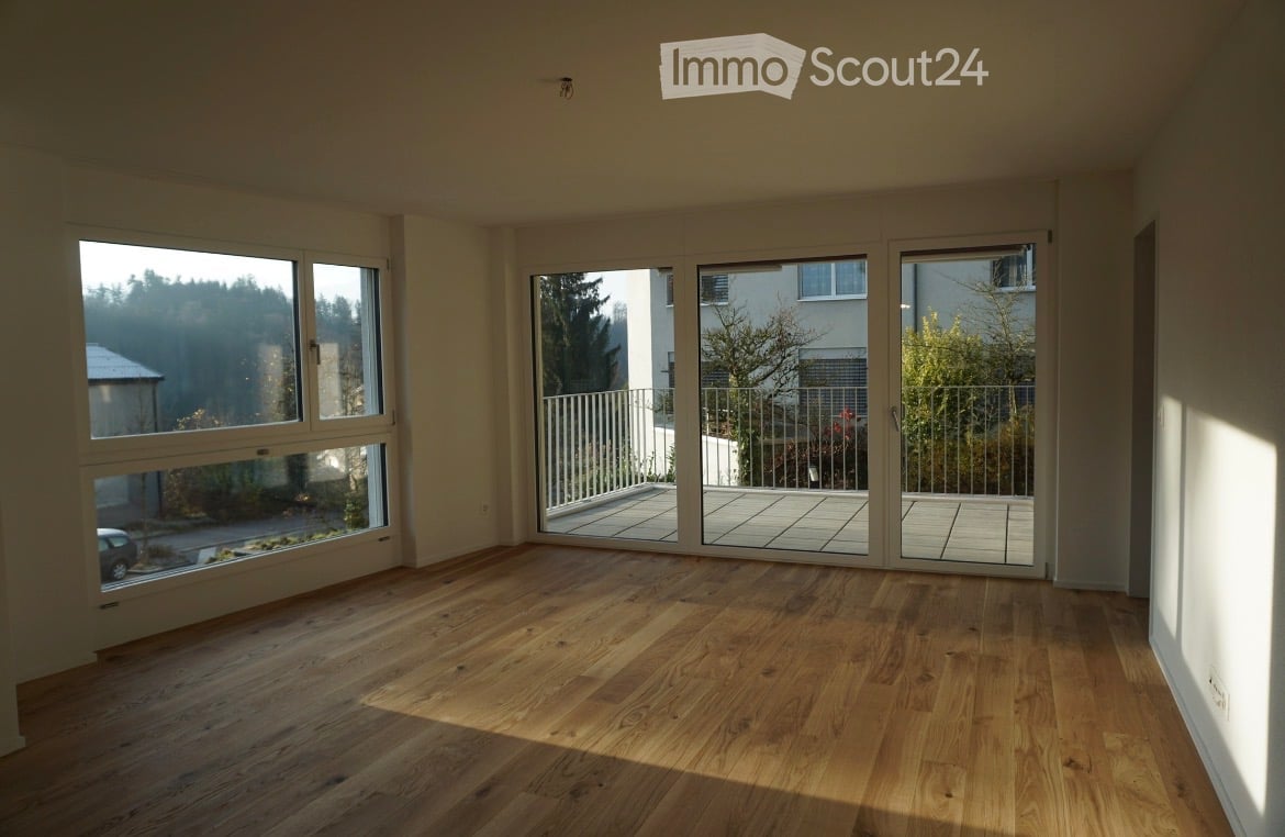 Empty room with wooden flooring, large windows, and sliding glass doors leading to a balcony.