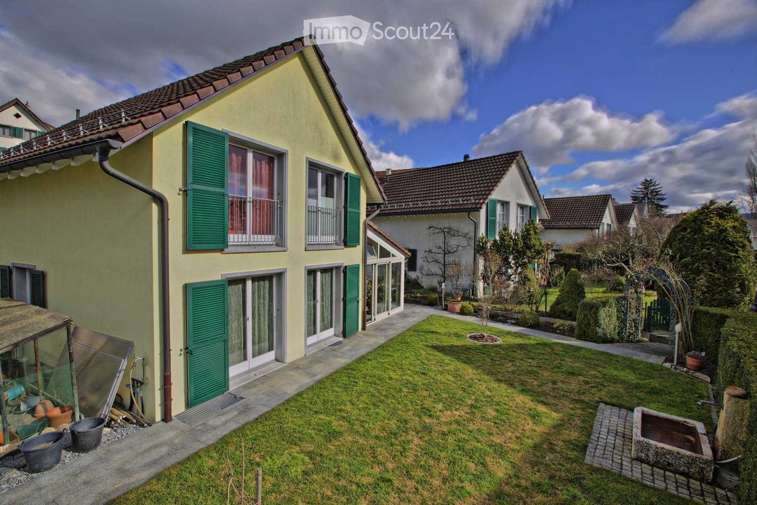 Two-story house with yellow exterior, green shutters, and multiple windows. The house features a well-maintained lawn, a small garden area with potted plants, and a paved pathway leading to the entrance.