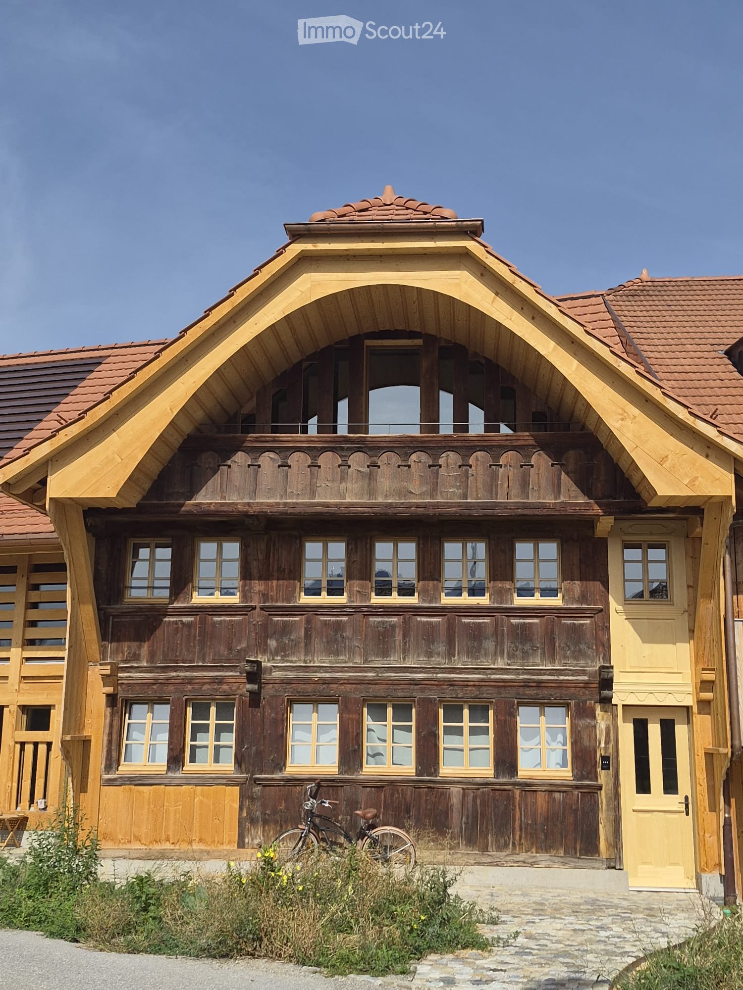Traditional wooden house with a steep, ornate roof and multiple windows, featuring a balcony and a bicycle parked in front.