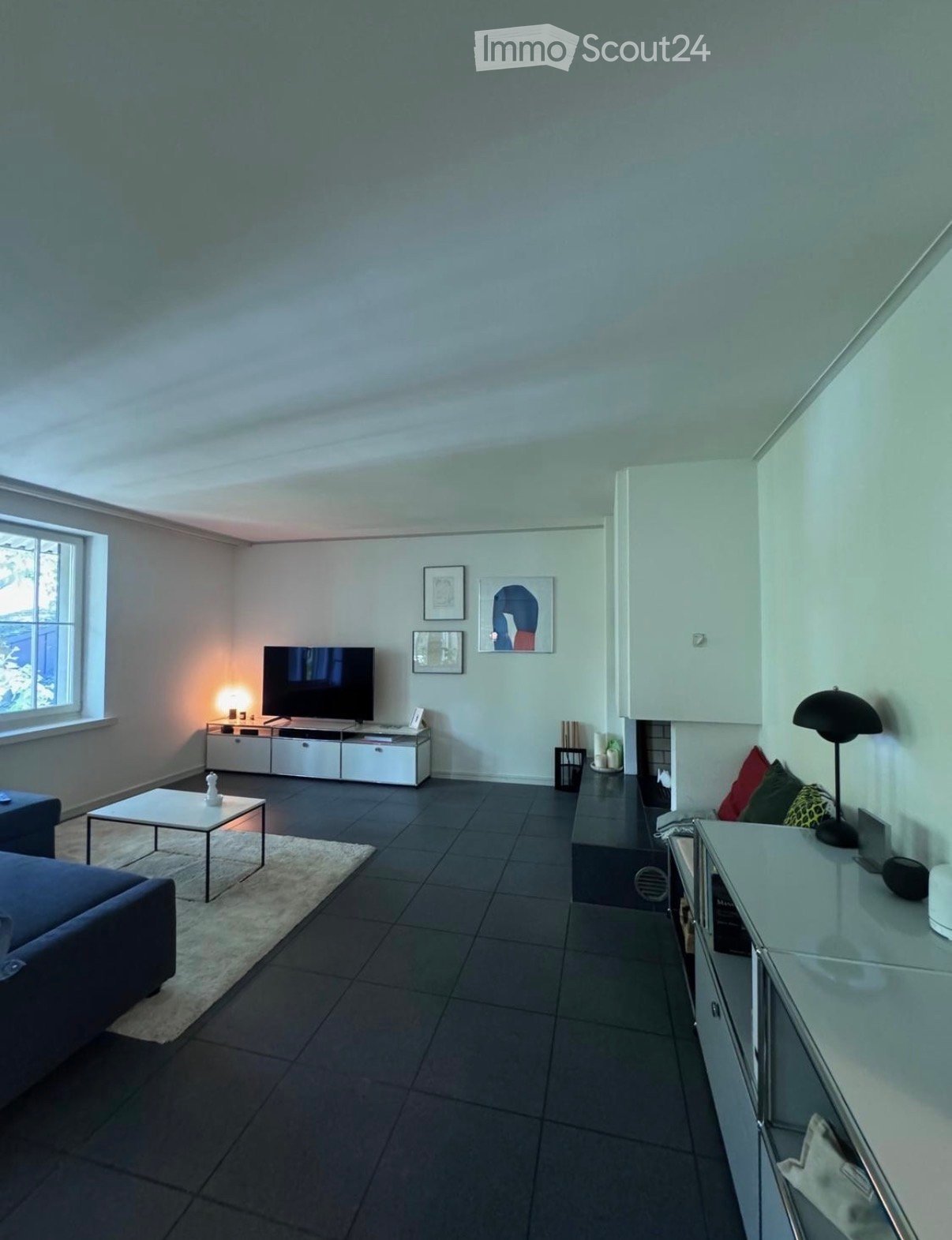 Modern living room with dark tiled flooring, a grey sofa, a white coffee table, a television on a white cabinet, and decorative picture frames on the wall.