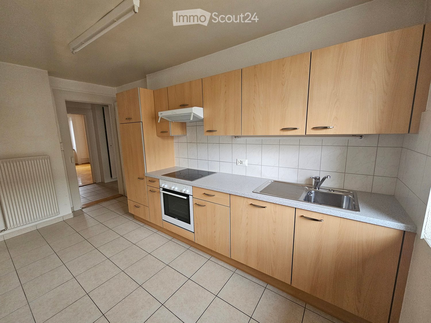 Modern kitchen with light wooden cabinets, a built-in oven, a sink with a faucet, and a range hood above the stove. The countertops are light-colored, and the floor is tiled.