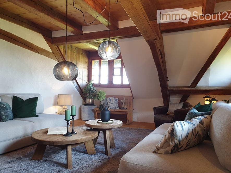 Cozy living room with exposed wooden beams, white walls, and large windows. Features a grey sofa, wooden coffee table, floor lamp, and decorative bench with plants.