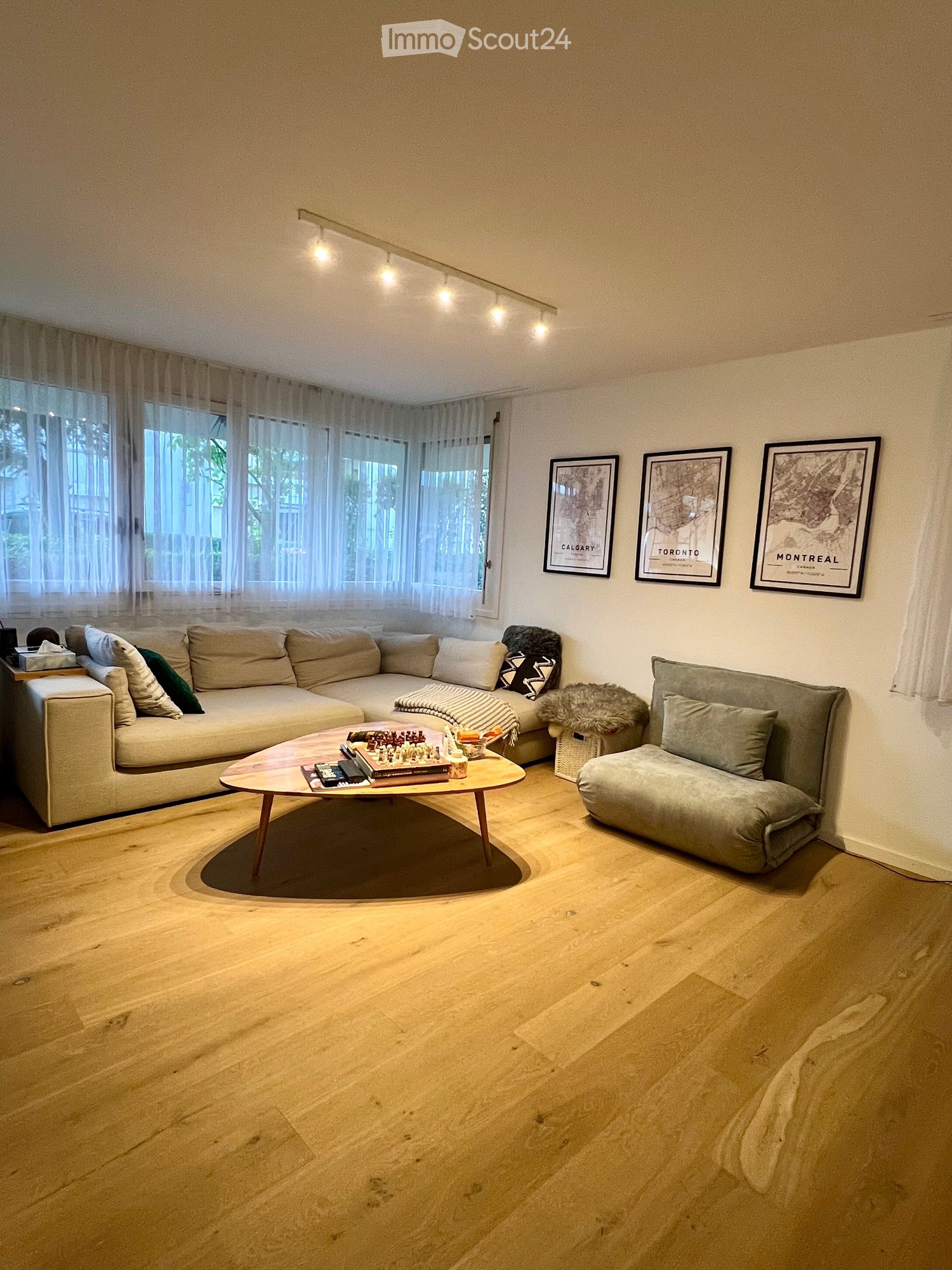 Modern living room with light wood flooring, large window with sheer curtains, sectional sofa, coffee table, armchair, and framed artwork on the wall.