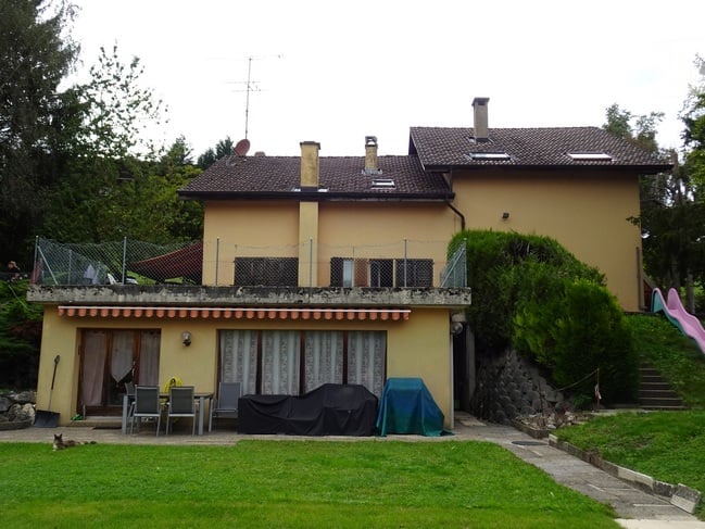 Two-story house with a tiled roof, multiple chimneys, and a covered balcony area. The front yard includes a patio with chairs and a table, and a small garden with a slide.