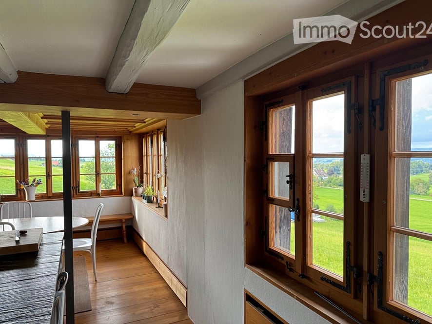 Bright, airy room with wooden floors and large windows overlooking a green field. Features a dining table with chairs, a bench, and several vases with flowers on the windowsill.