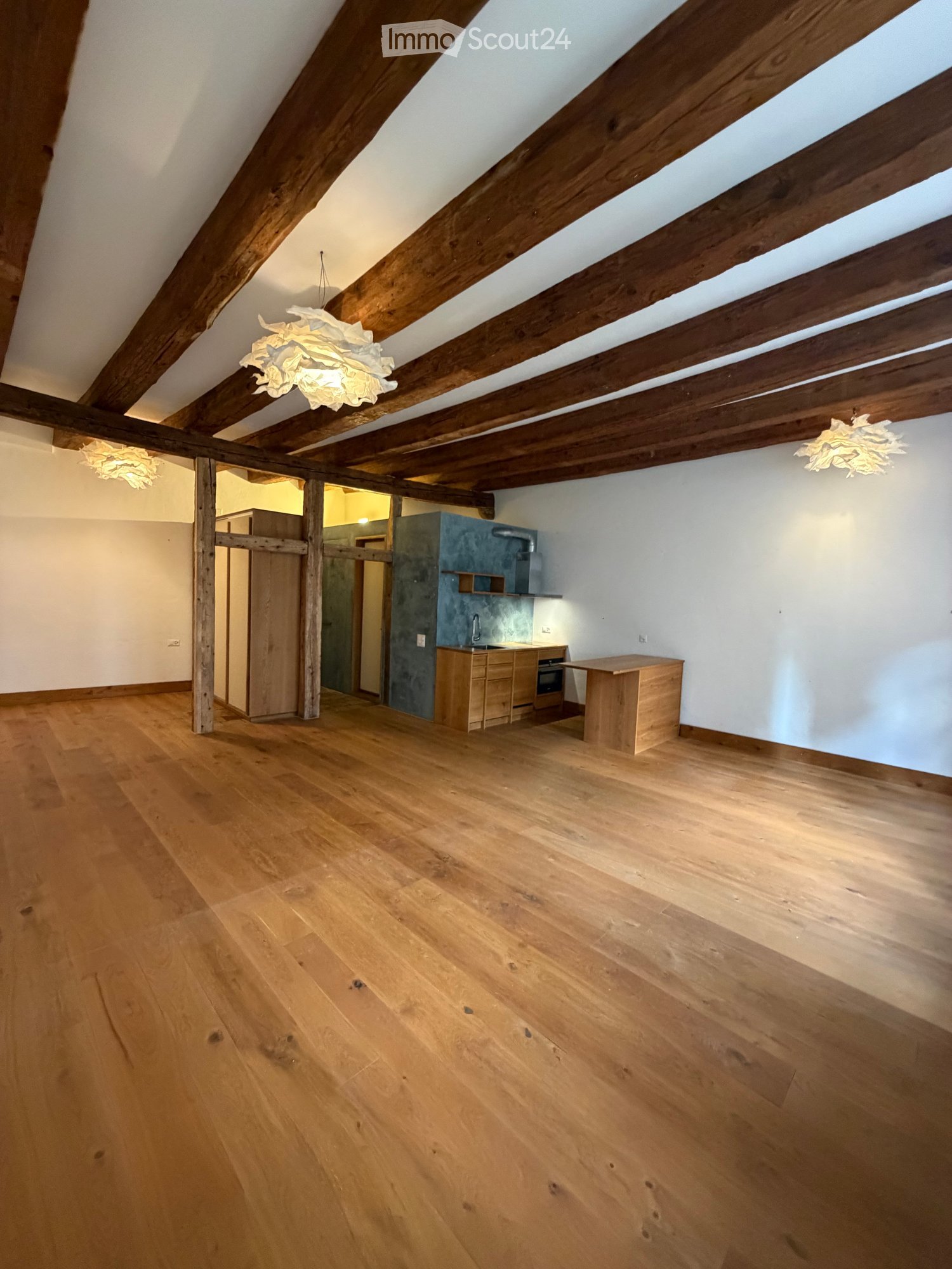 Open-plan attic space with wooden beams, exposed wooden floor, and a small kitchen area with cabinets, sink, and oven. A wooden table is placed near the kitchen.