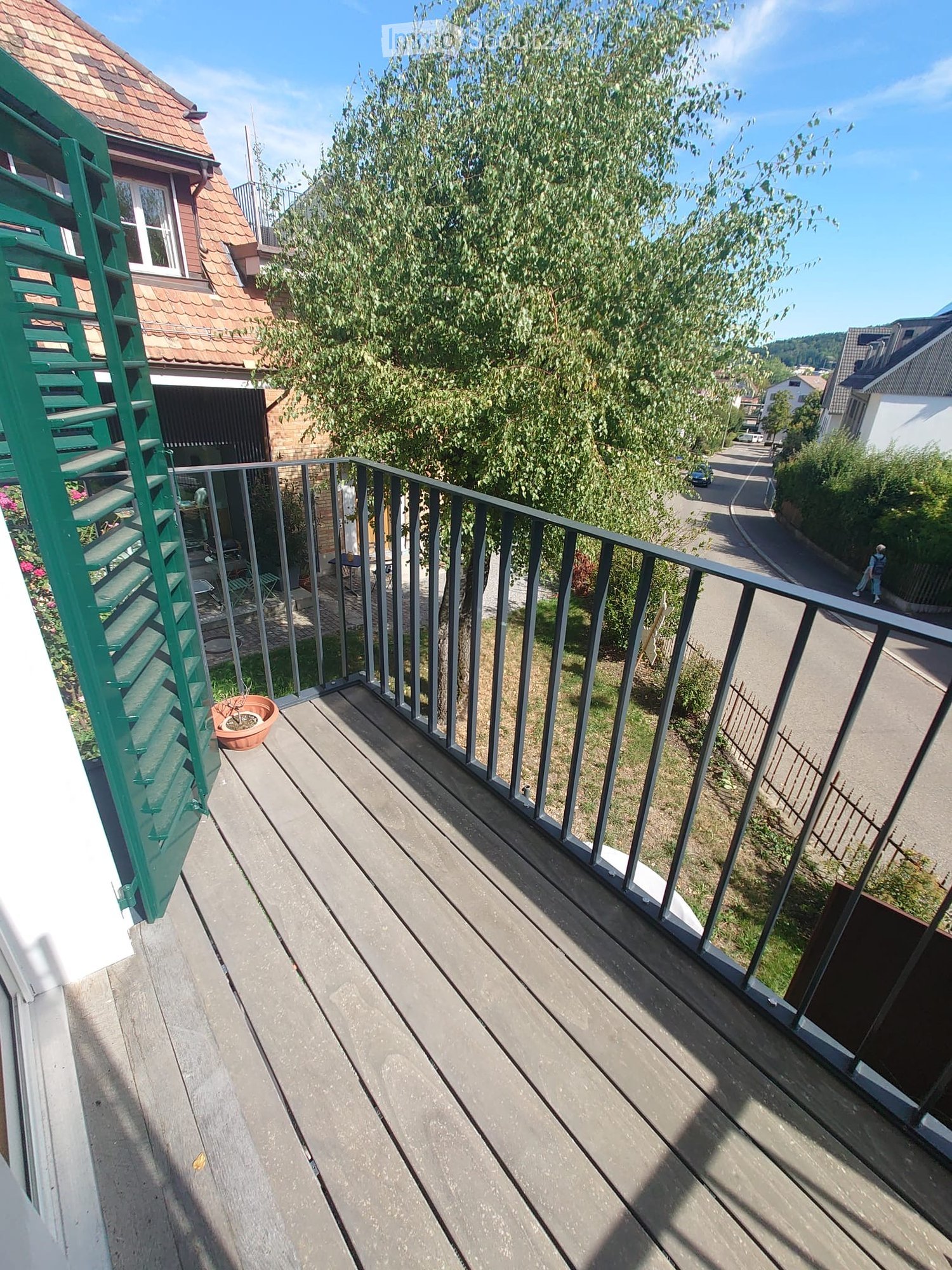 Balcony with wooden decking, metal railing, and a potted plant visible. The view shows a residential street with houses and trees.