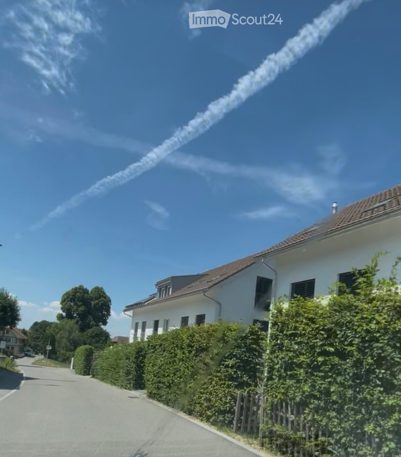 A two-story white house with a brown roof, surrounded by greenery and a clear blue sky with contrails from an aircraft.