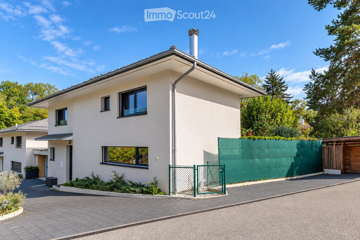 Modern two-story house with white exterior, large windows, and a green fence surrounding the property.