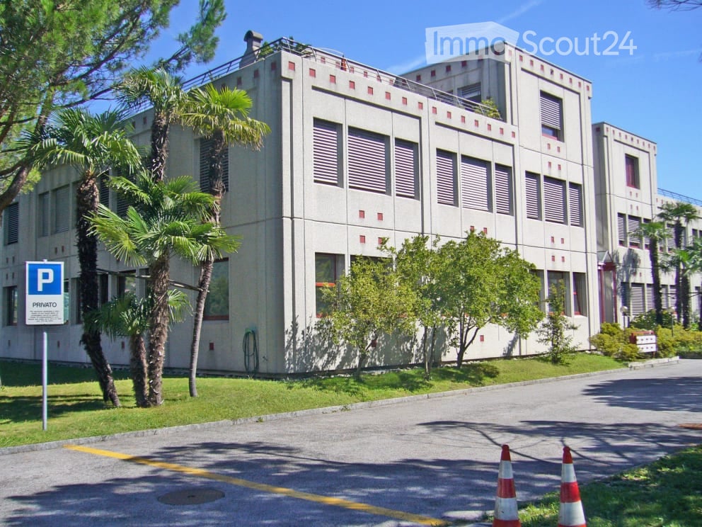 Modern multi-story building with a clean, geometric facade, surrounded by palm trees and greenery. A parking sign is visible in the foreground.