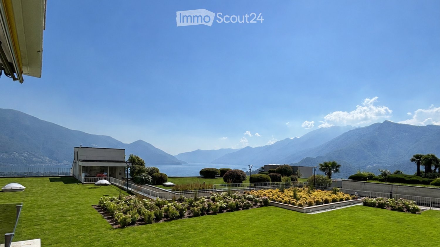 Well-maintained garden with neatly trimmed hedges, flower beds, and a small building, overlooking a scenic lake and mountain range.