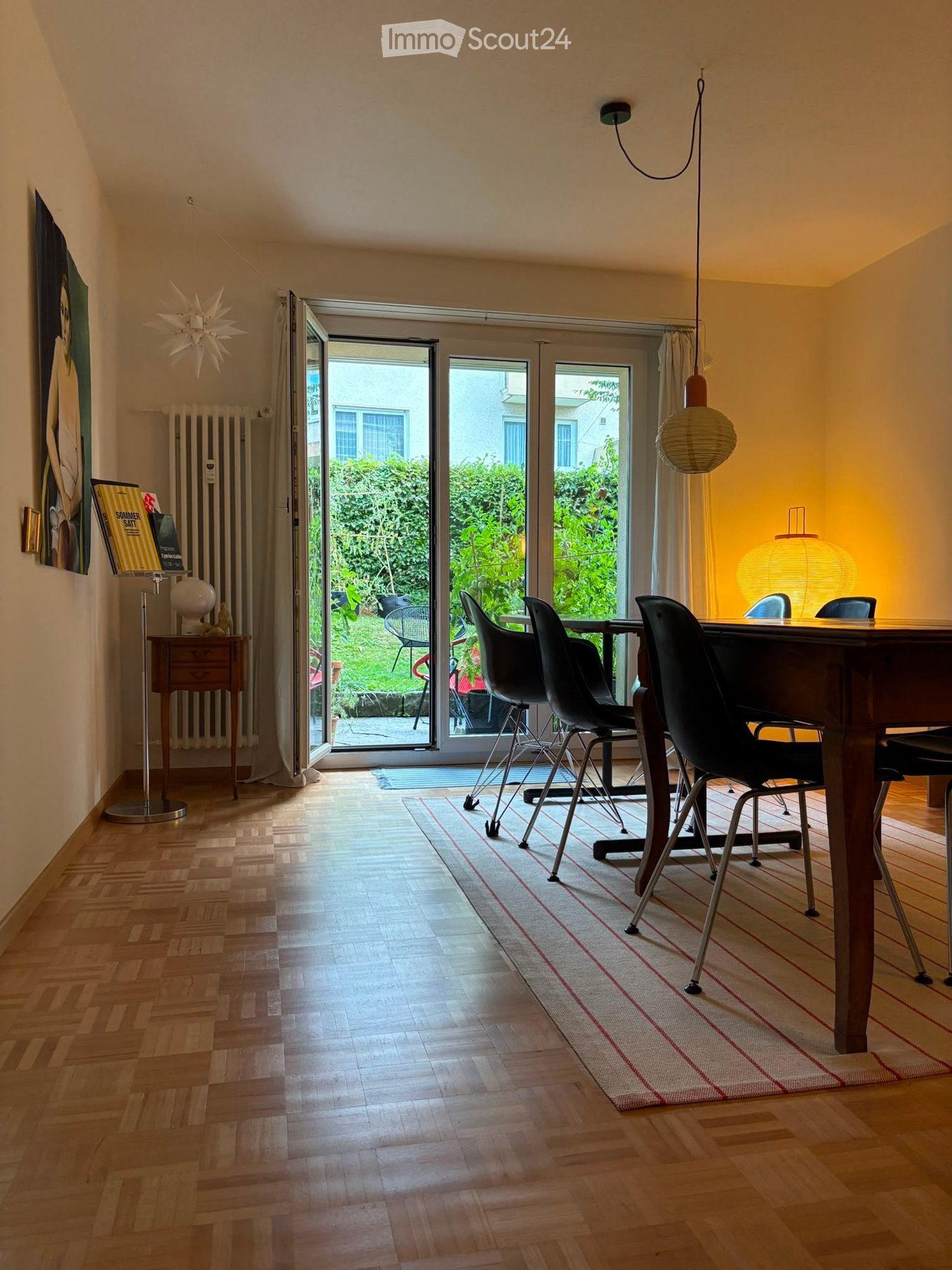 Bright dining area with wooden flooring, large sliding glass doors opening to a garden, a wooden dining table with black chairs, a hanging lamp, a floor lamp, a radiator, a globe, and a painting on the wall.