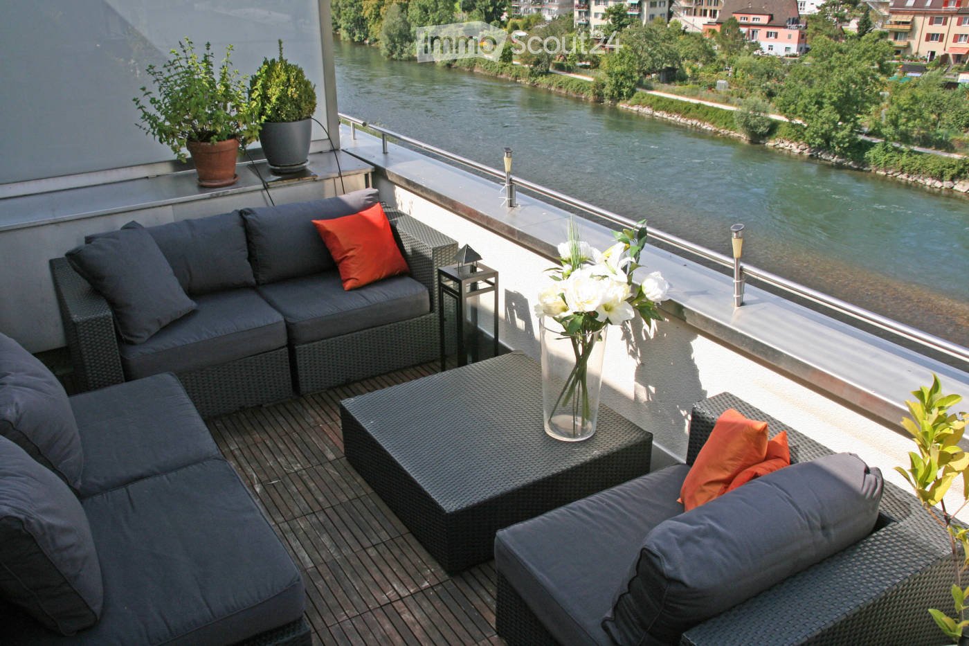 Balcony with a view of a river, featuring a grey wicker sofa, a black wicker coffee table, a vase with white flowers, and potted plants.