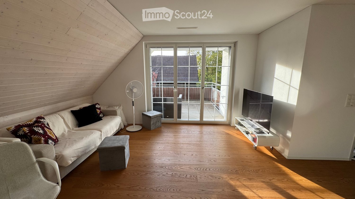 Bright living room with wooden flooring, a white sofa with patterned cushions, a fan, a TV on a stand, and access to a balcony.