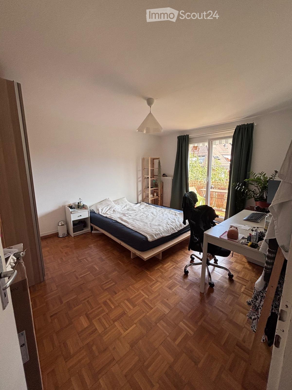 Cozy bedroom with wooden flooring, a single bed with white bedding, a nightstand, a desk with a laptop, a chair, and a shelf with books and a plant. Large window with dark curtains and a view of the garden.
