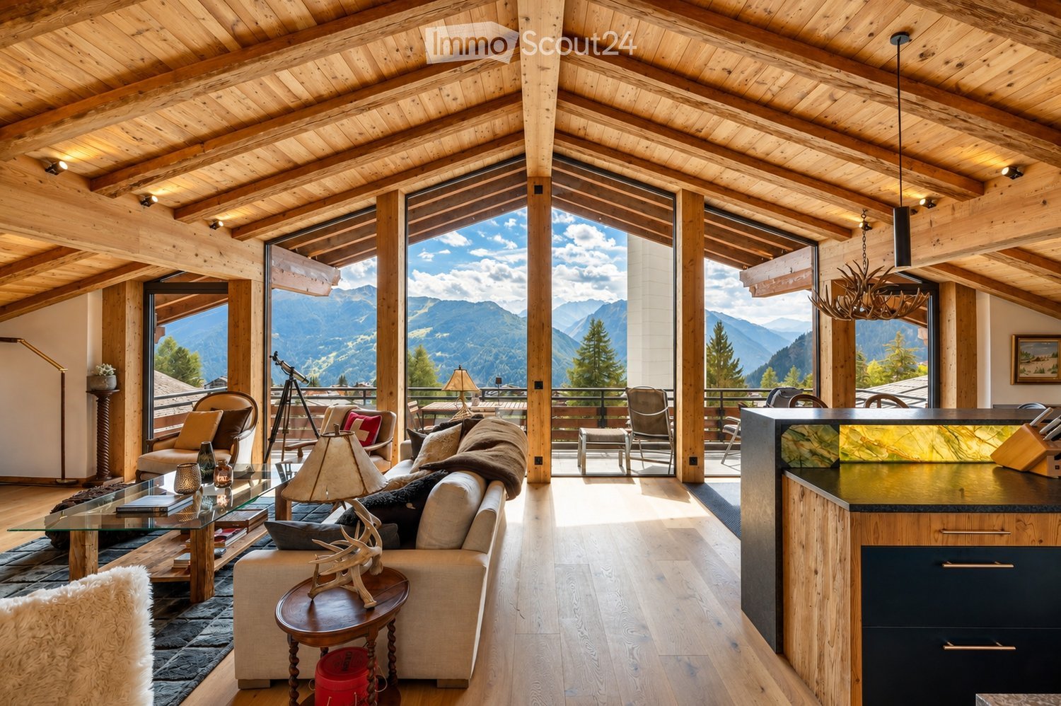 A spacious, well-lit living room with exposed wooden beams and large windows offering a scenic mountain view. The room features a cream-colored sofa, a glass coffee table, a wooden side table with a decorative deer head, and a modern kitchen island. Addit