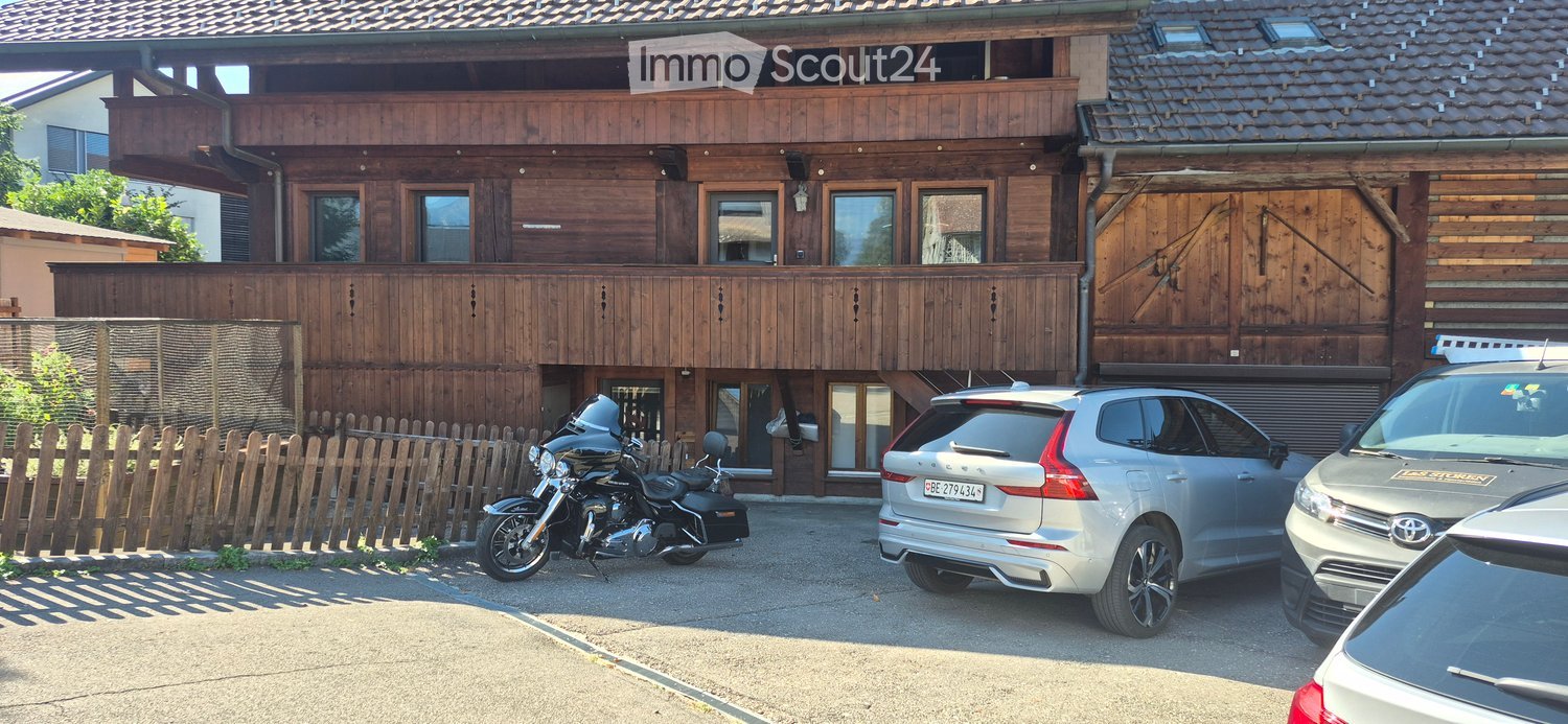 A two-story wooden house with a balcony, garage, and several parked vehicles including a motorcycle and cars.