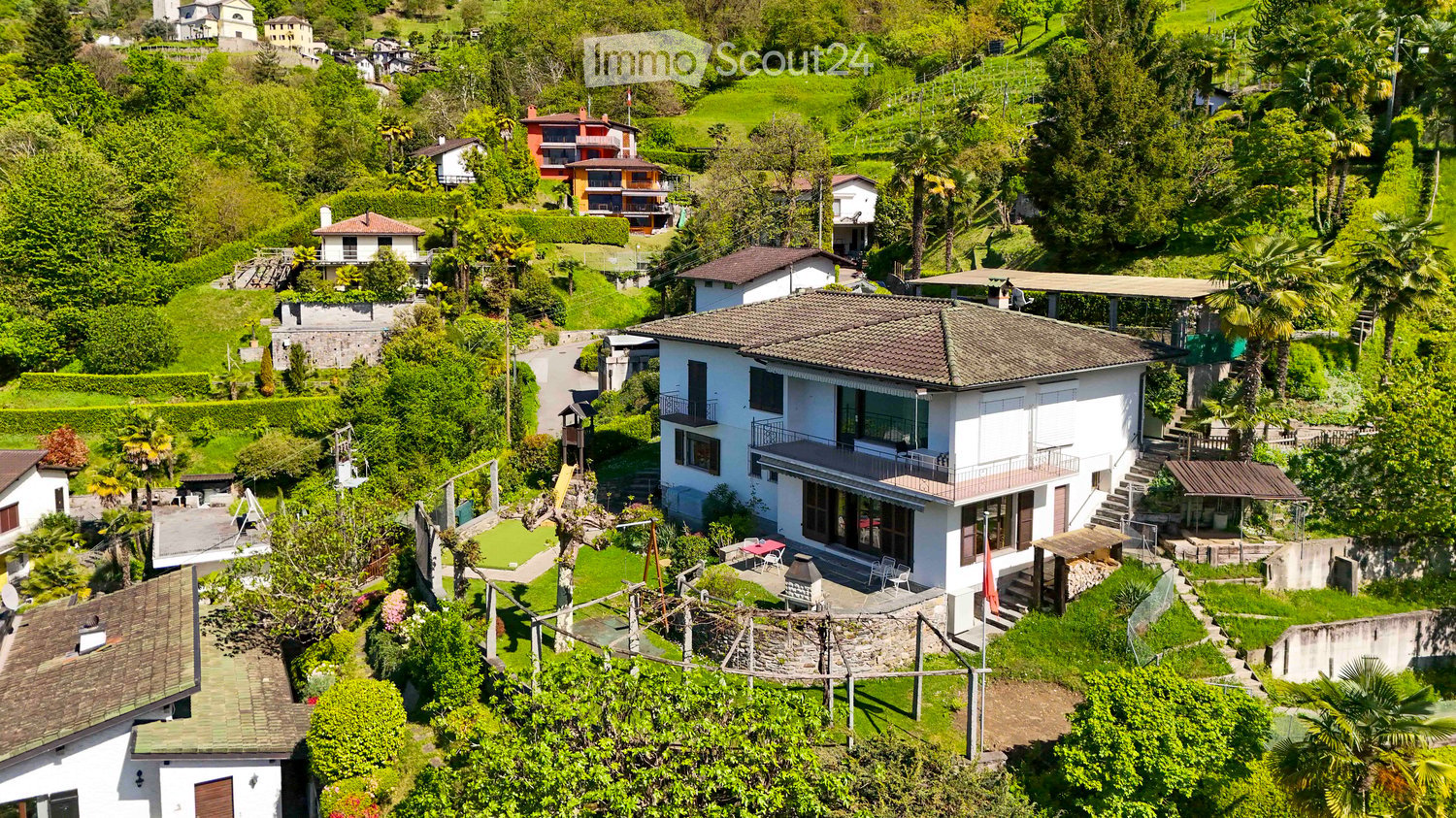 Aerial view of a residential area with multiple houses surrounded by lush greenery and trees. The houses have varied architectural styles and are situated on a hilly terrain.