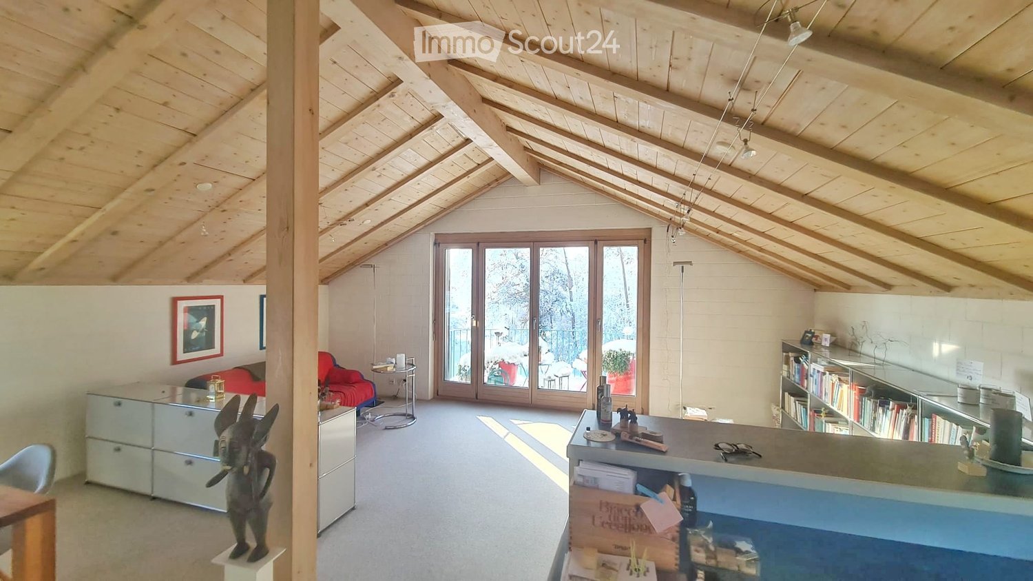 Bright, open attic space with wooden beams, large windows, and a balcony. Features a red sofa, glass table, bookshelf, and decorative items.